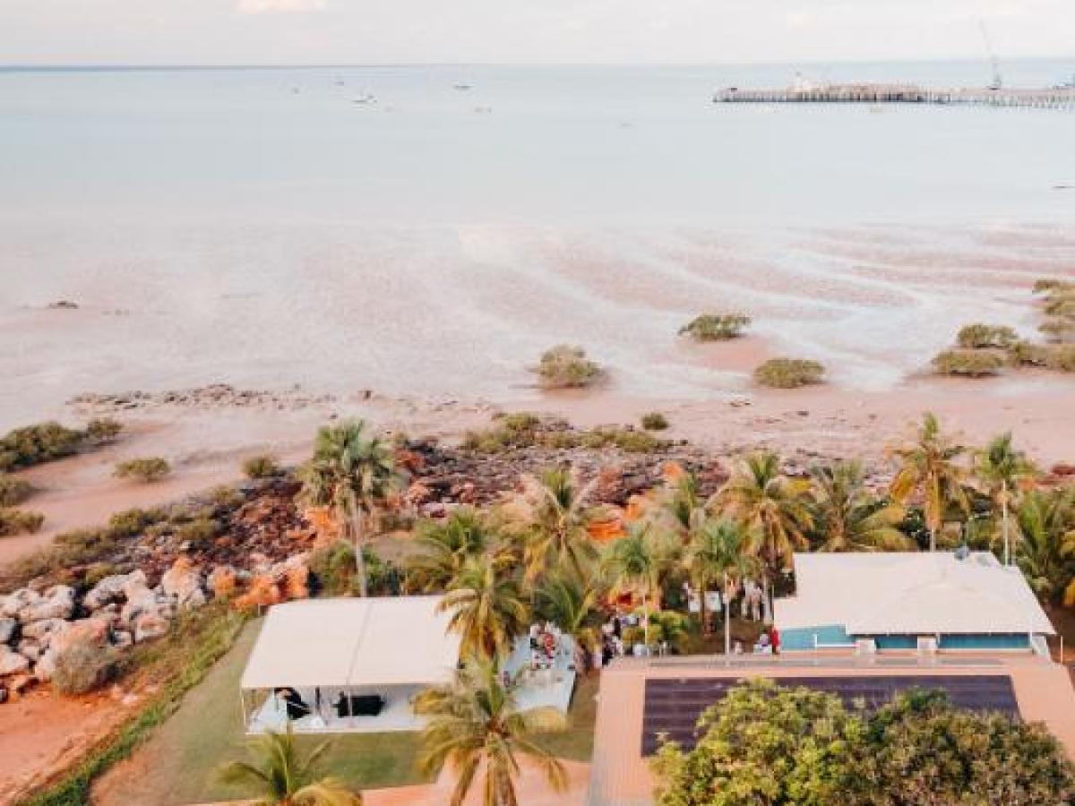 Aerial view of beach, palm trees, and houses near calm sea under cloudy sky.
