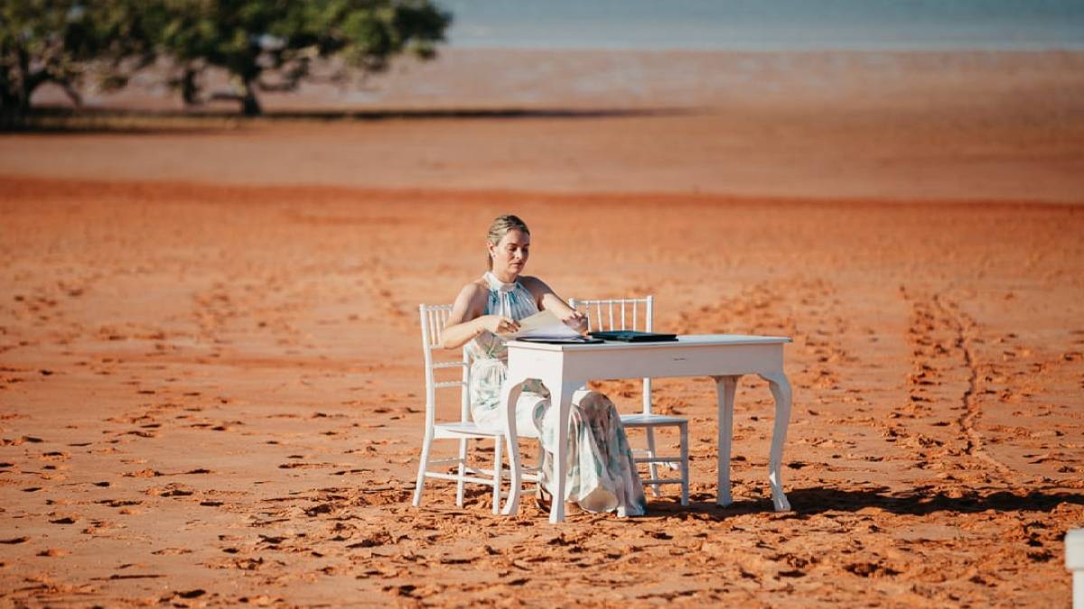 Person seated at a white table on sandy beach with trees and water in background.