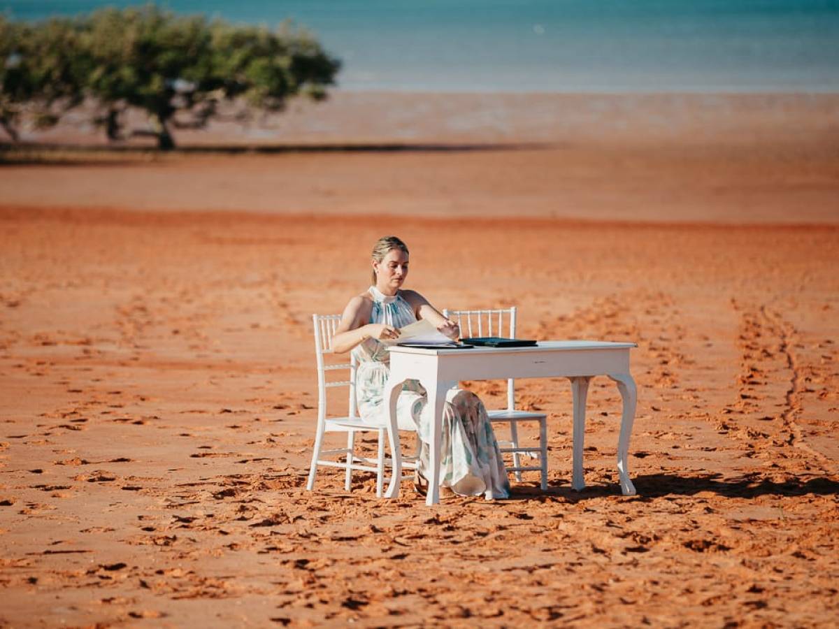 Person seated at a white table on sandy beach with trees and water in background.