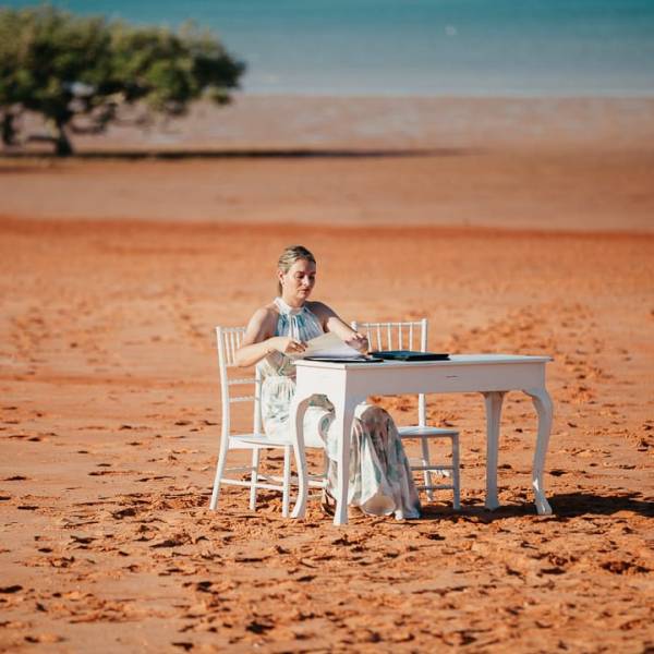 Person seated at a white table on sandy beach with trees and water in background.