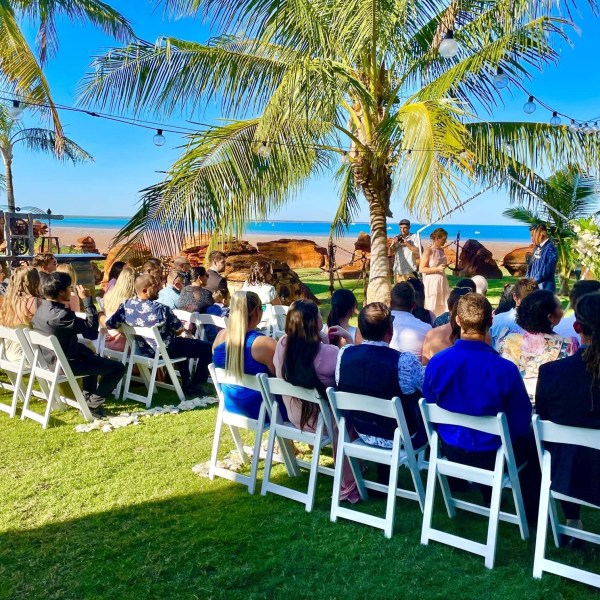 Outdoor wedding ceremony on a beach with palm trees and guests seated in white chairs.