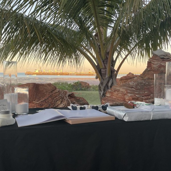 Outdoor table with candles, gifts, and a book under palm tree near a beach at sunset.