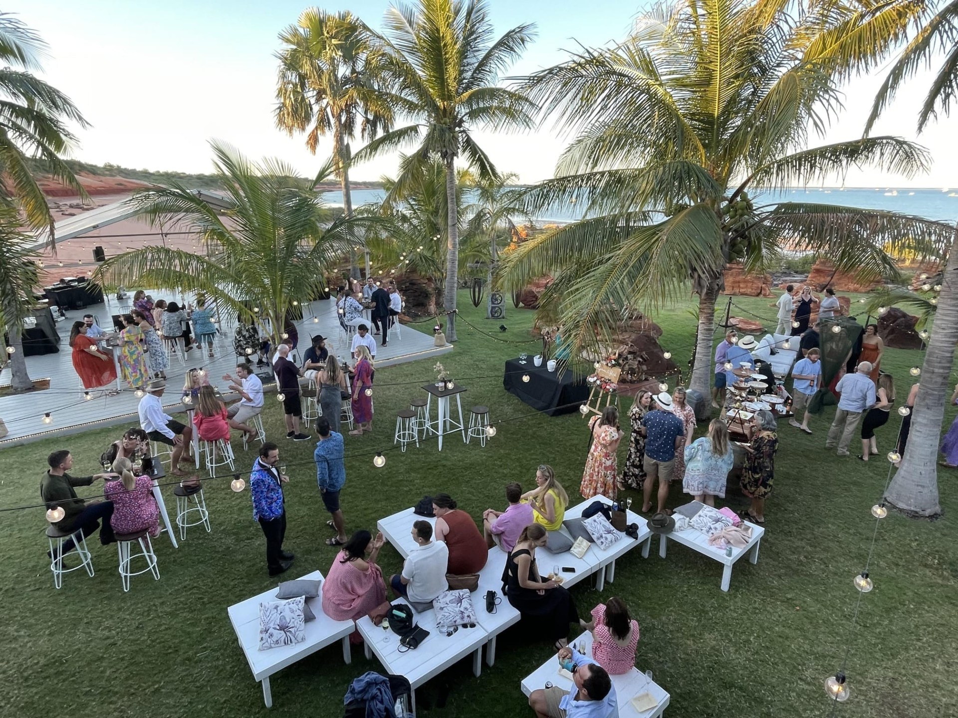 Outdoor party with people mingling under palm trees, near a stage and tables, by the ocean.