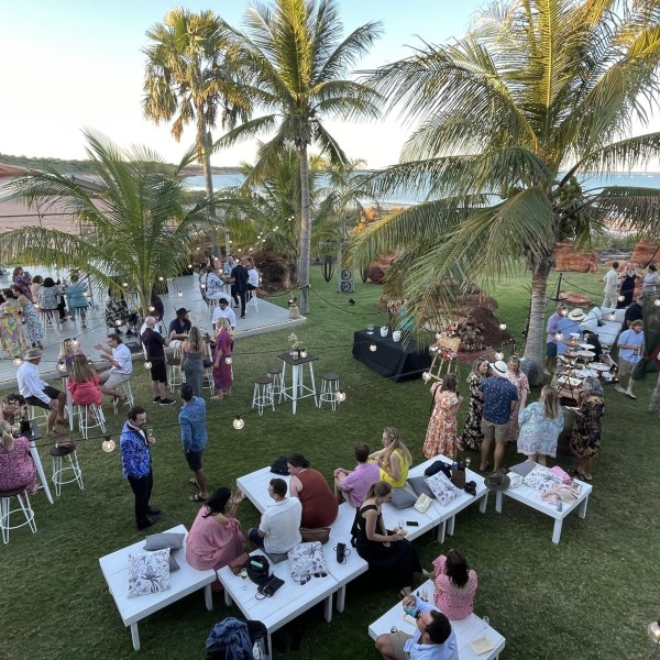 Outdoor party with people mingling under palm trees, near a stage and tables, by the ocean.