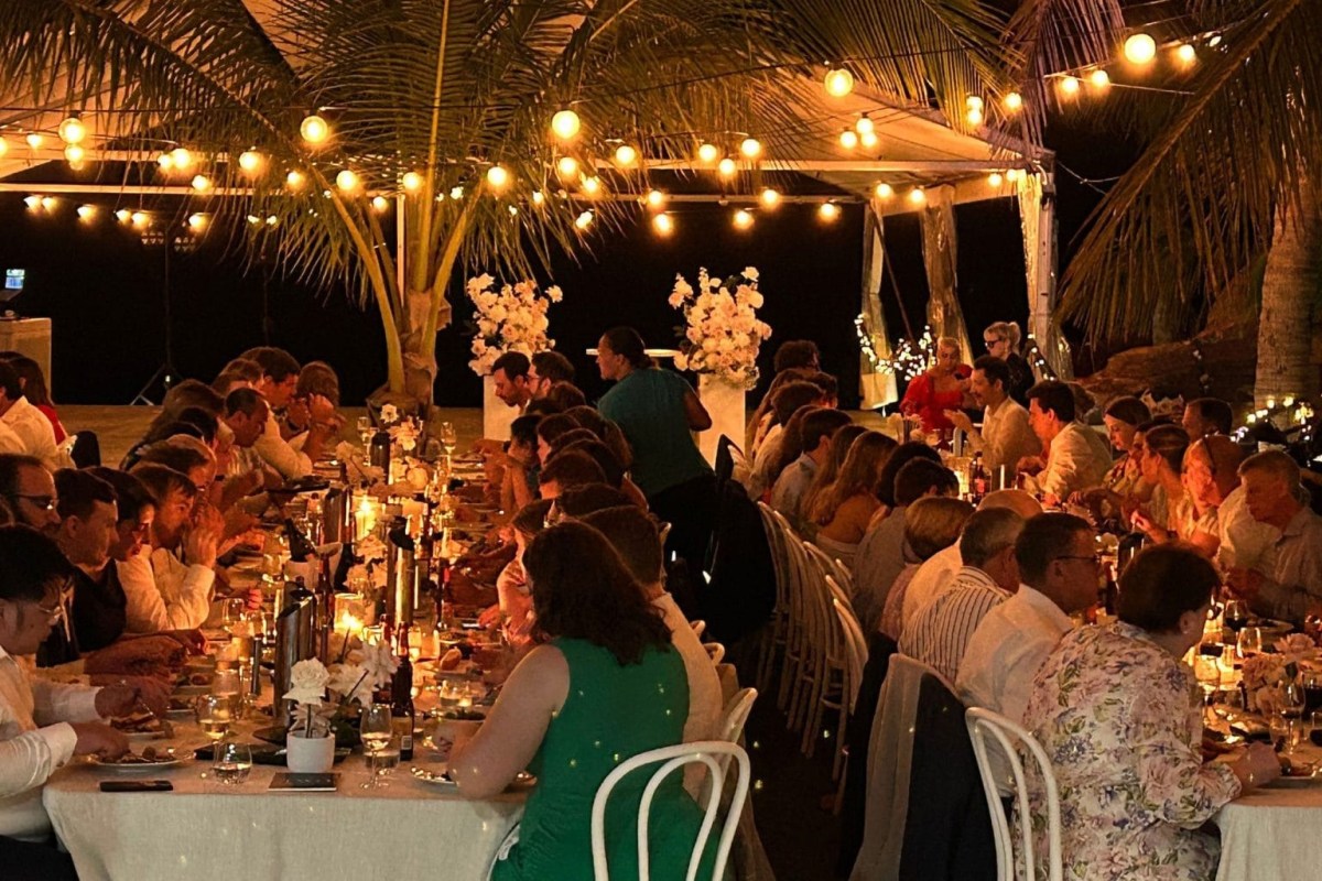 Large outdoor dinner party with guests seated under string lights and palm trees.