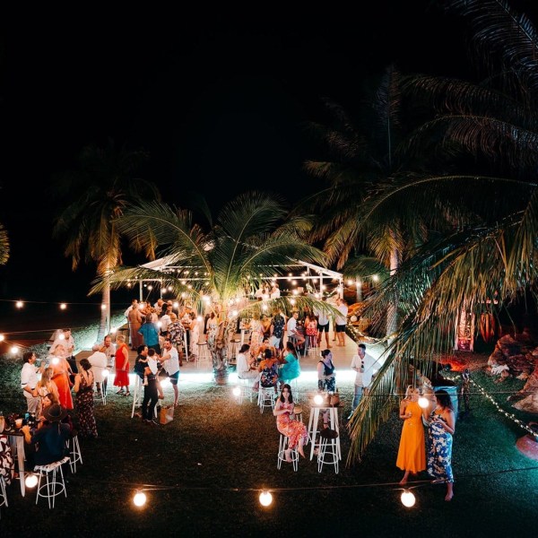 Outdoor evening party with string lights, guests mingling under palm trees.