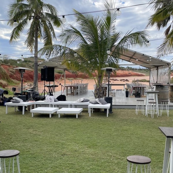 Outdoor event setup with tables, chairs, palm trees, and string lights under a cloudy sky.