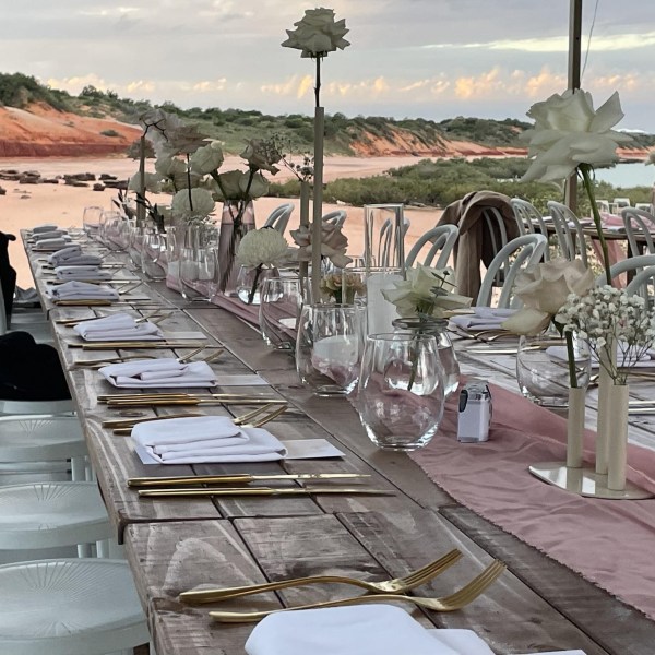 Outdoor dining table setup with white chairs, beach view in the background.
