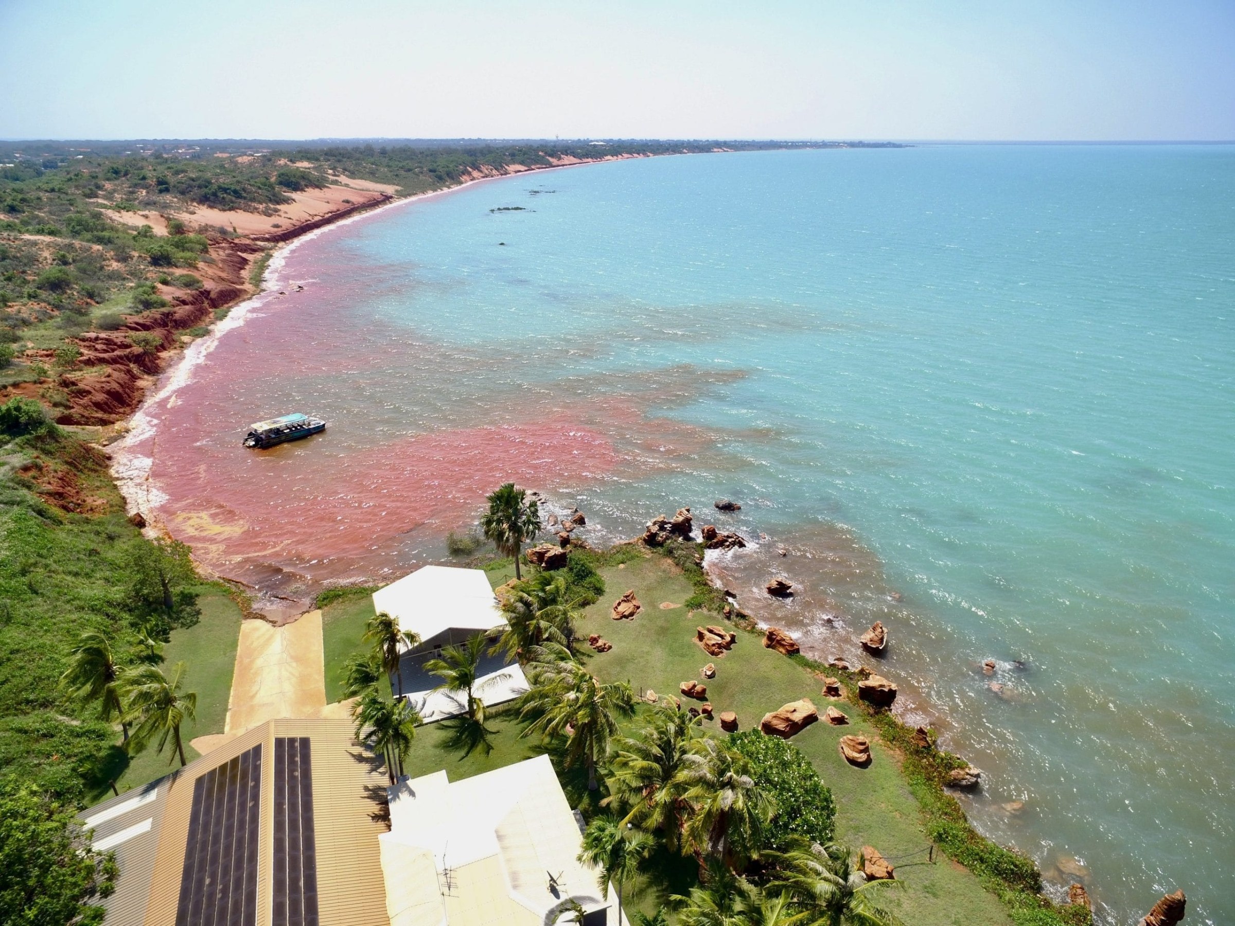 Aerial view of a coastal area with red cliffs, green grass, palm trees, and turquoise water.