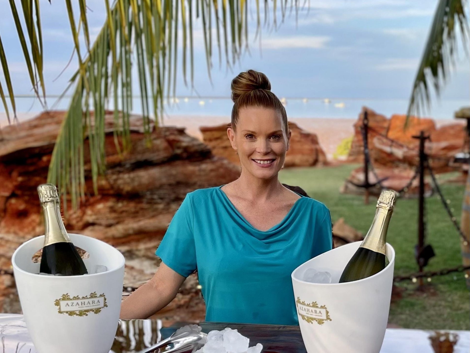 Woman in teal shirt behind table with two champagne bottles in ice buckets near beach.