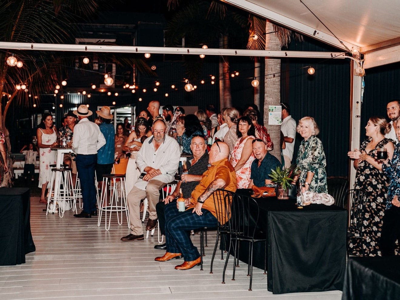 A lively outdoor gathering with people sitting and standing under string lights.