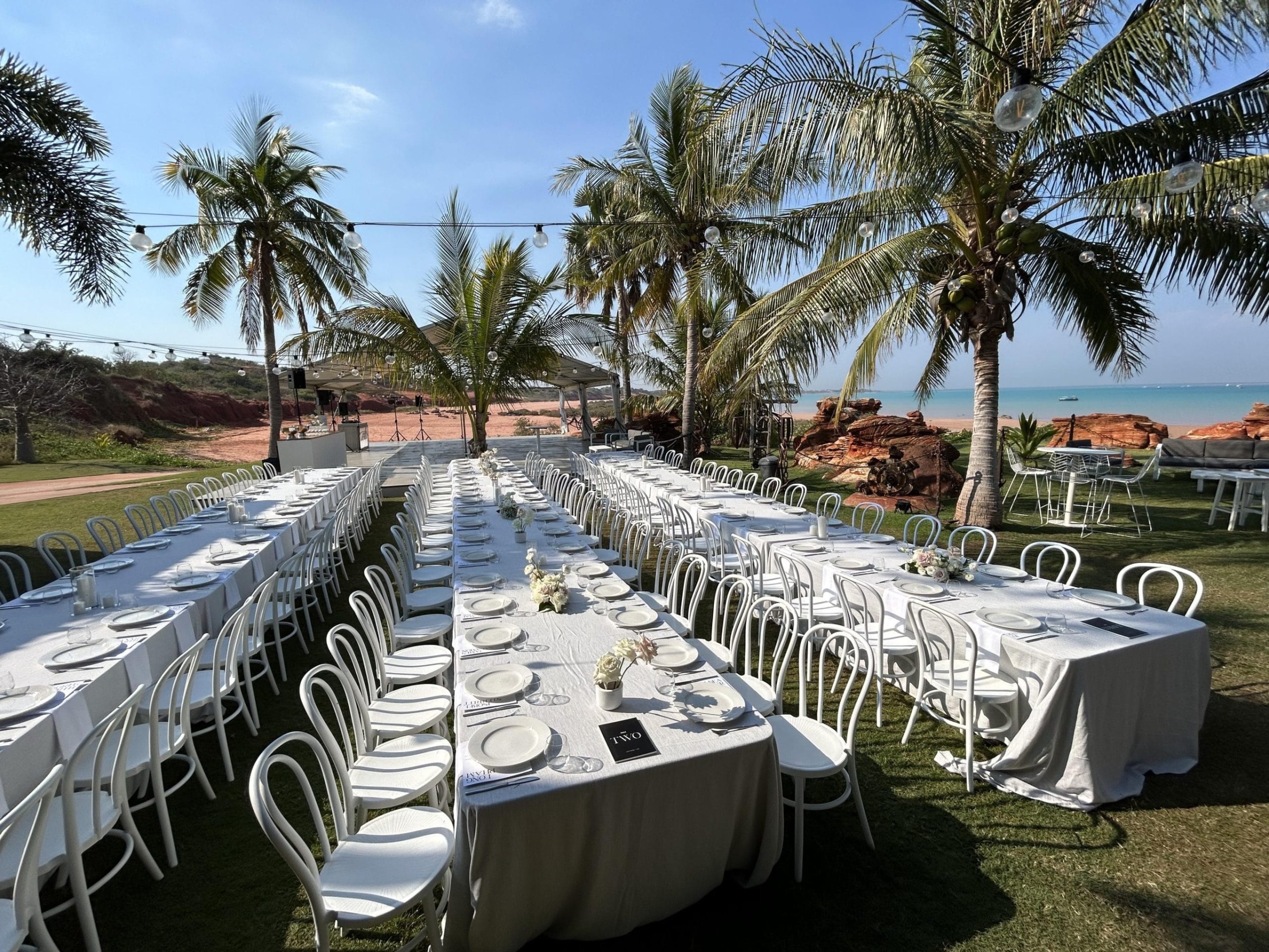 Outdoor tables set for dining under palm trees by the ocean.