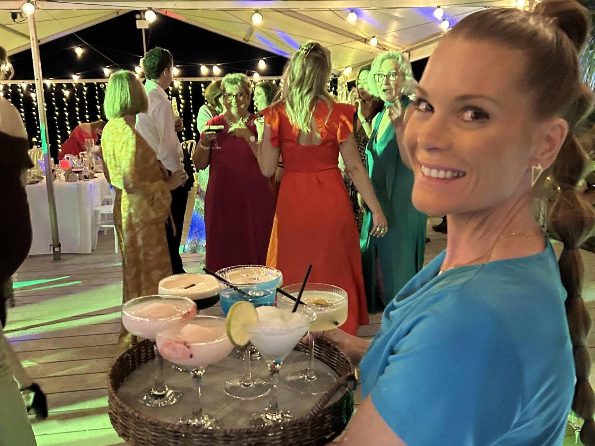 Smiling woman in blue dress holds tray with cocktails at a party under a tent.
