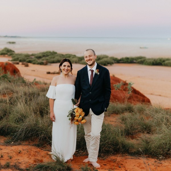Bride and groom standing on a beach with grass and colorful sky in the background.