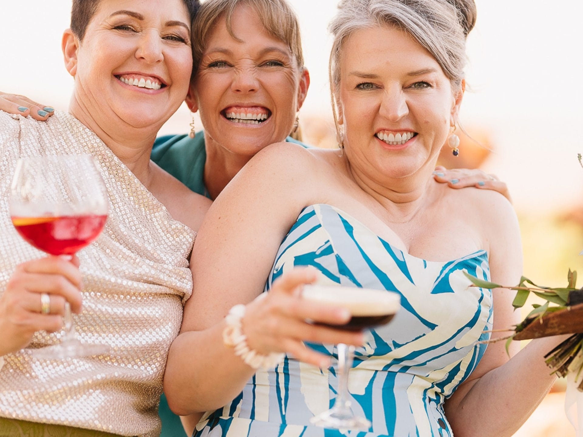 Three women smiling and holding drinks outdoors in bright, patterned dresses.