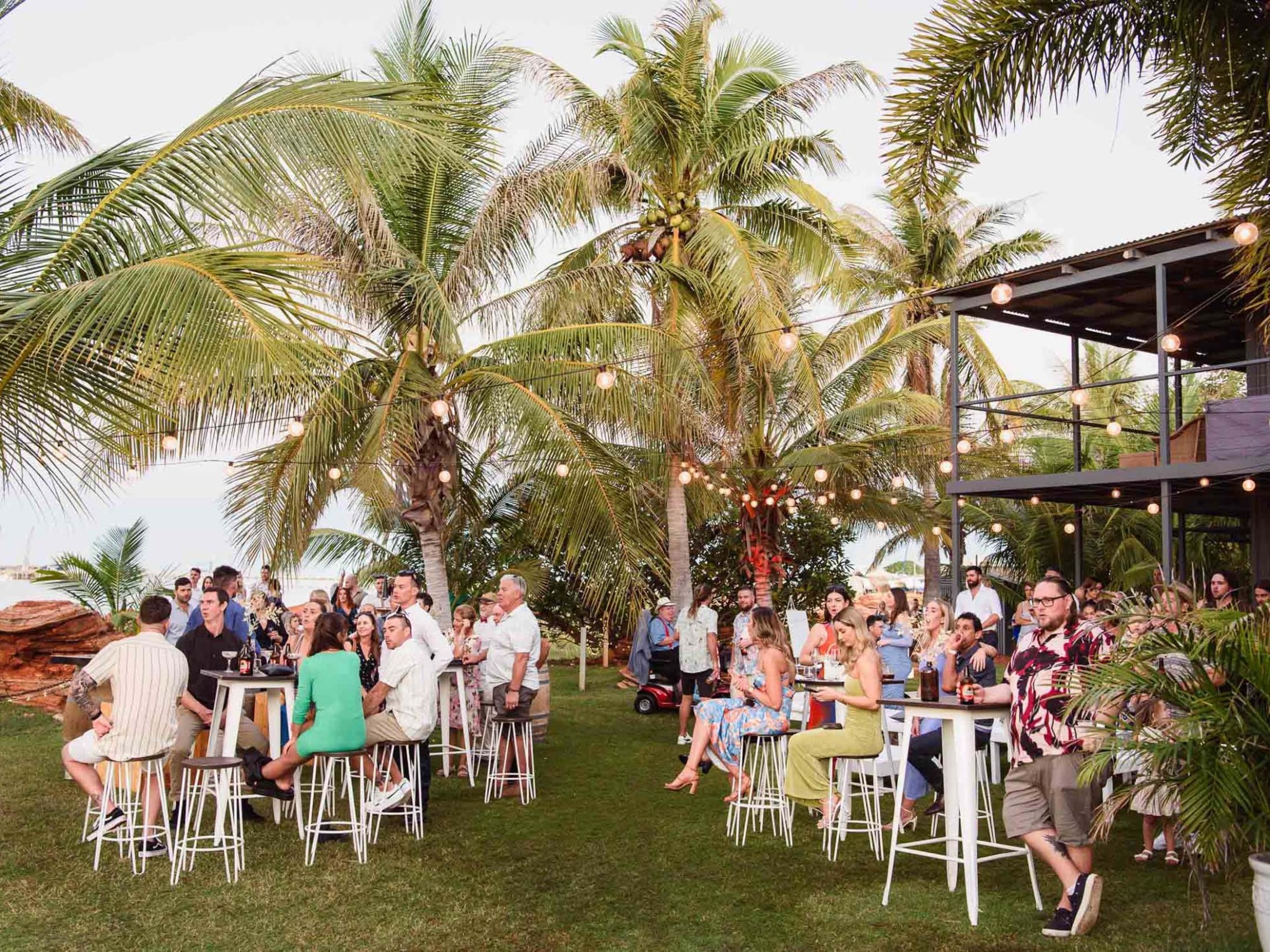 Outdoor party with people around tables under palm trees and string lights.