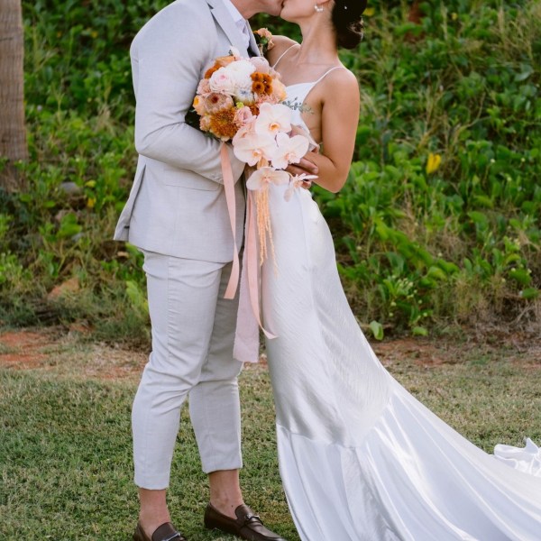 Couple in wedding attire kissing outdoors, bride holding bouquet, green background.