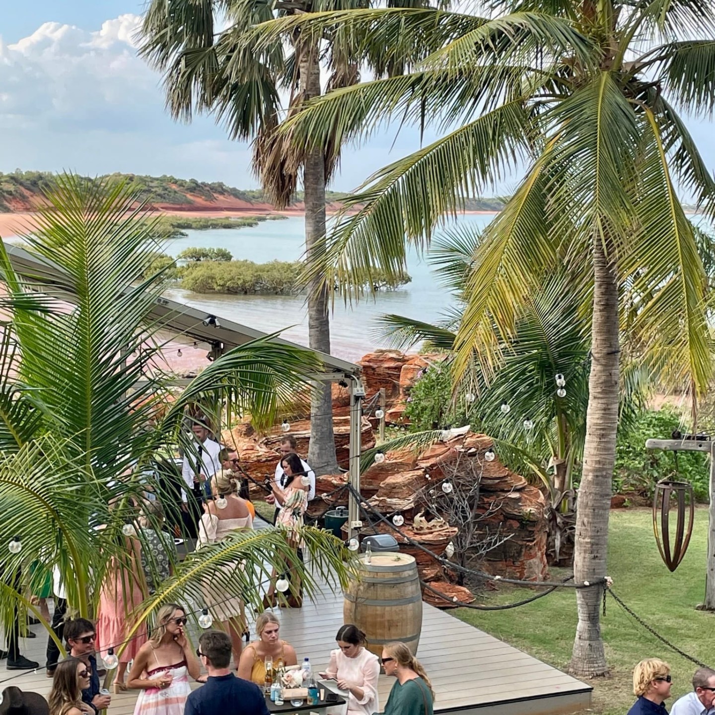 People socializing outdoors near beach and palm trees, with a coastal backdrop.