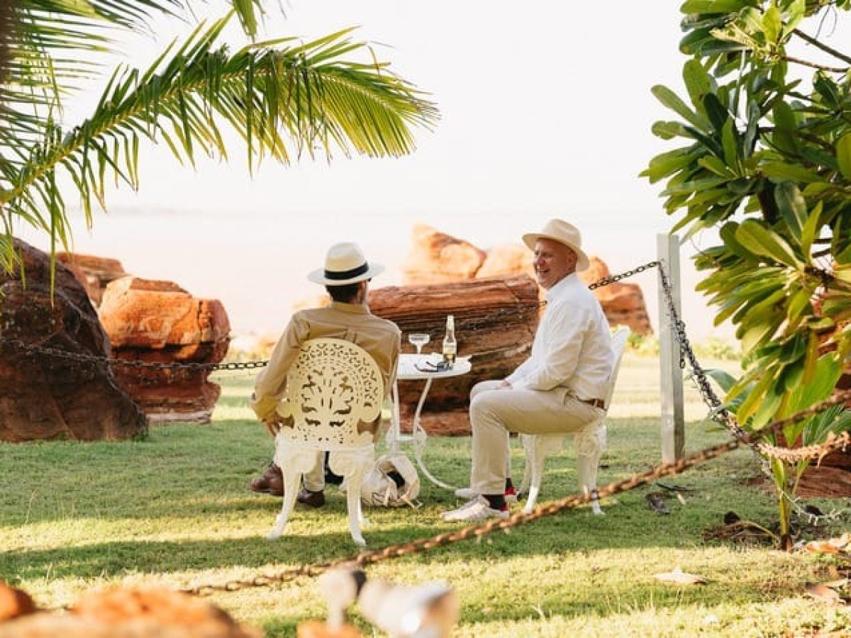 Two people in hats sit outdoors on white chairs near rocks, with a palm and sea in the background.