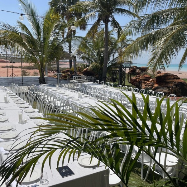 Outdoor event setup with long tables, white chairs, palm trees, and ocean view.