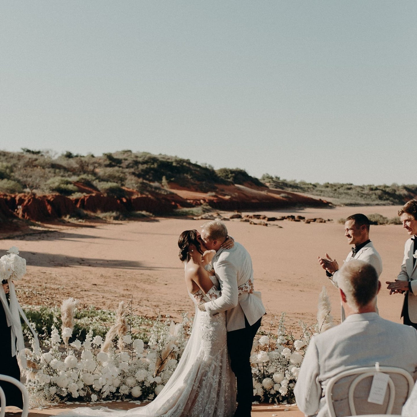 Couple kissing at outdoor wedding ceremony with guests seated and desert landscape background.