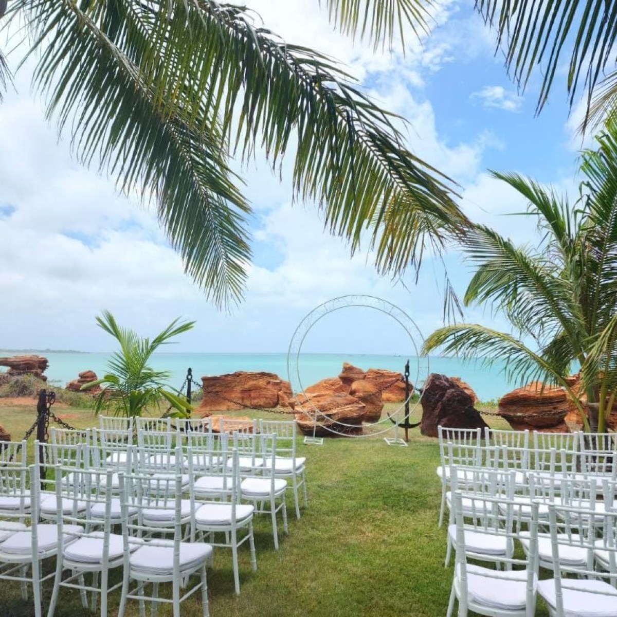 Outdoor beach wedding setup with white chairs, circular arch, and palm trees.