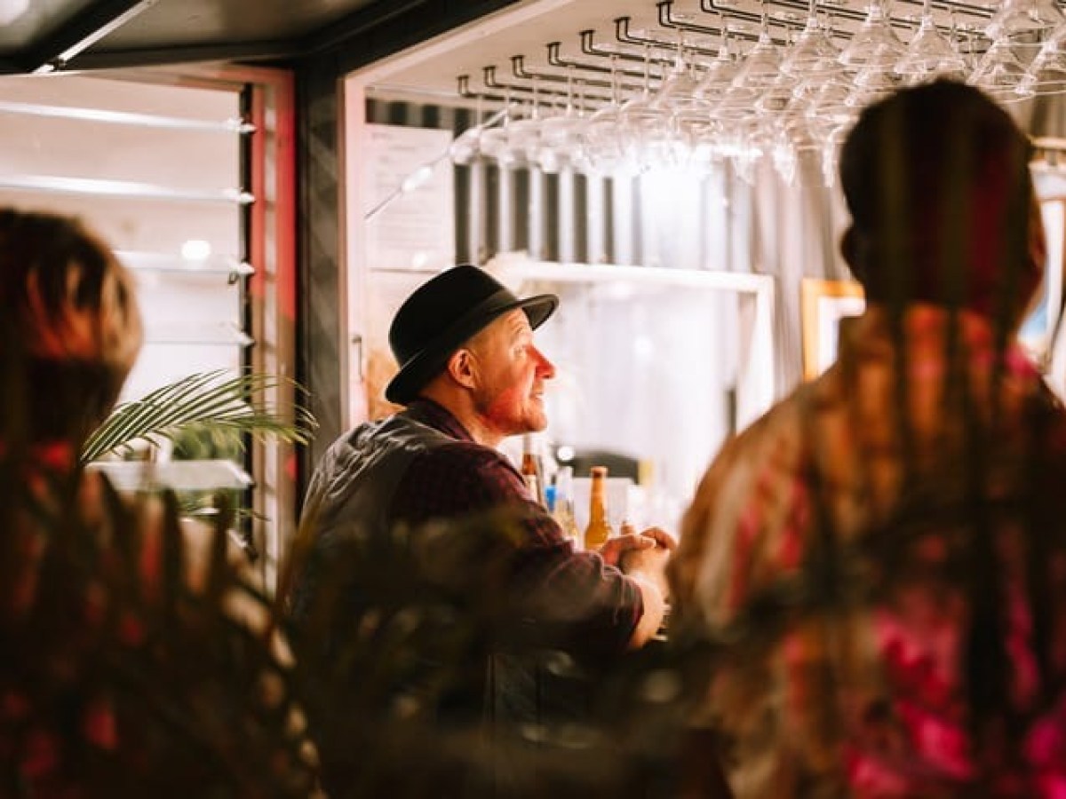 Man in hat smiling at a bar with people and plants around.