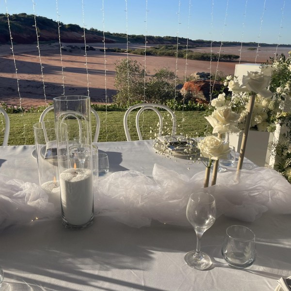 Elegant outdoor table setting with candles and white roses, overlooking a green landscape under clear skies.