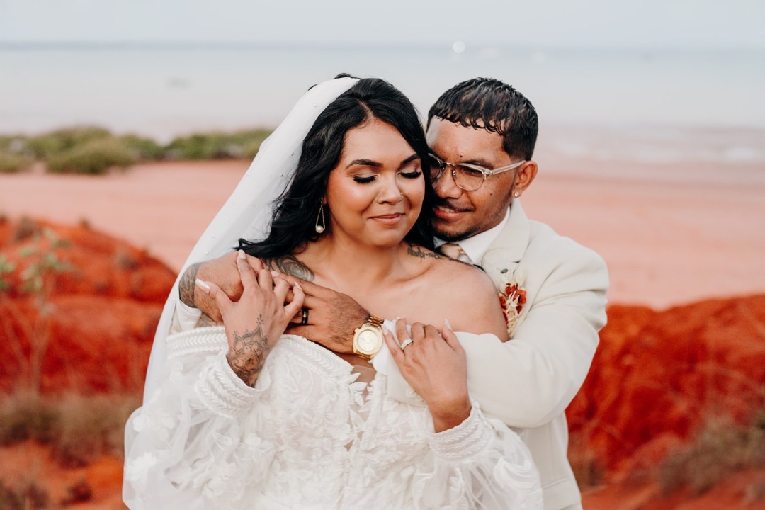 Bride and groom embrace in a desert setting with red earth and blue sky.