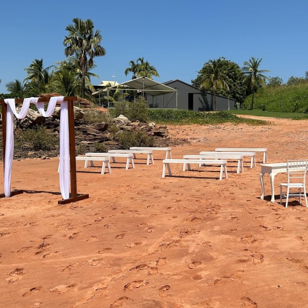 Beach wedding setup with wooden arch, white benches, and chairs under clear blue sky.