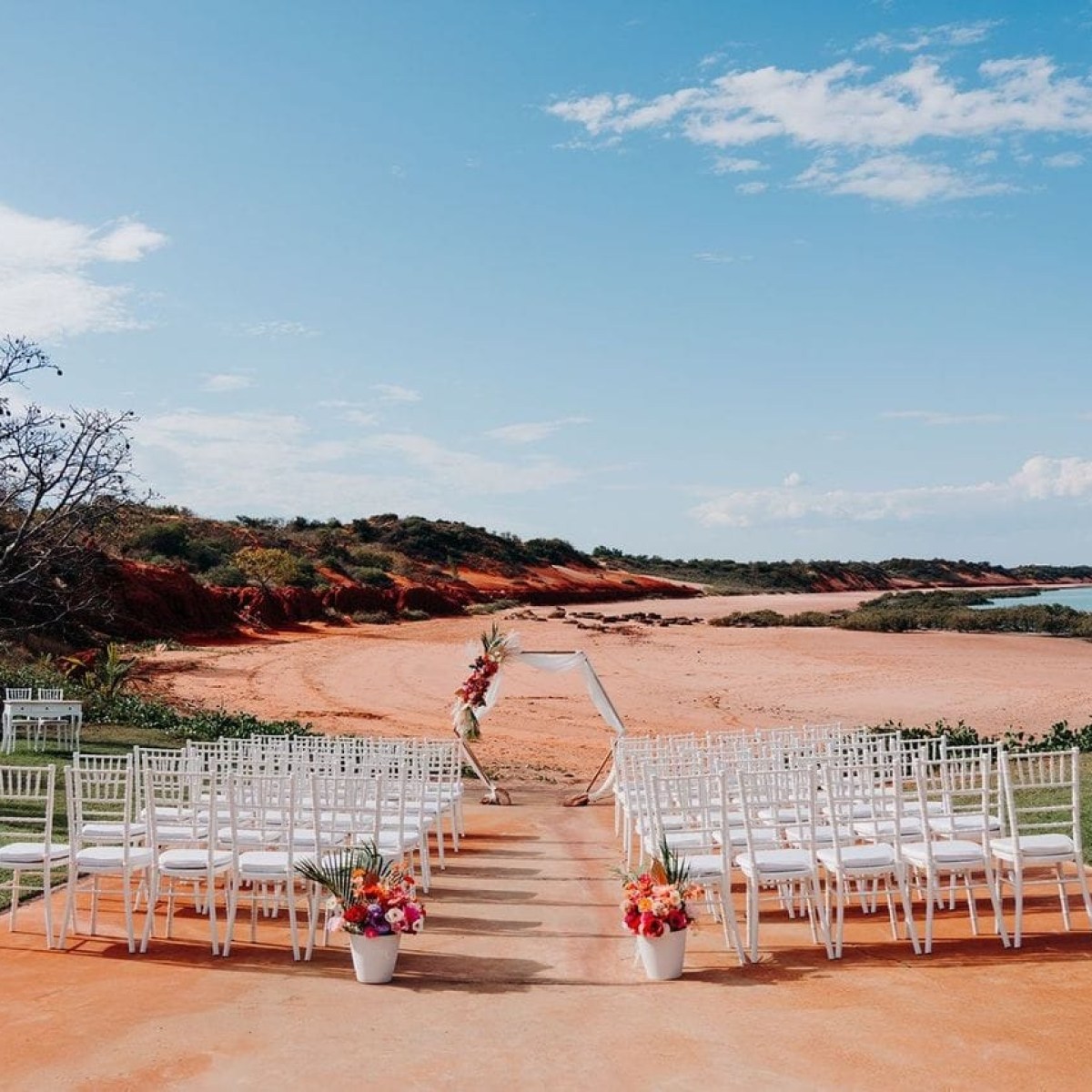 Outdoor wedding setup on a beach with white chairs and floral decorations under a clear sky.
