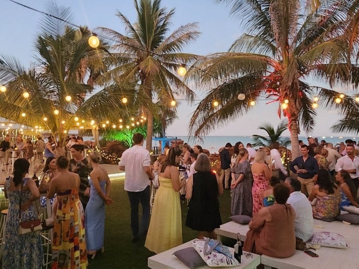 Outdoor beach party with string lights, palm trees, and people socializing in evening setting.