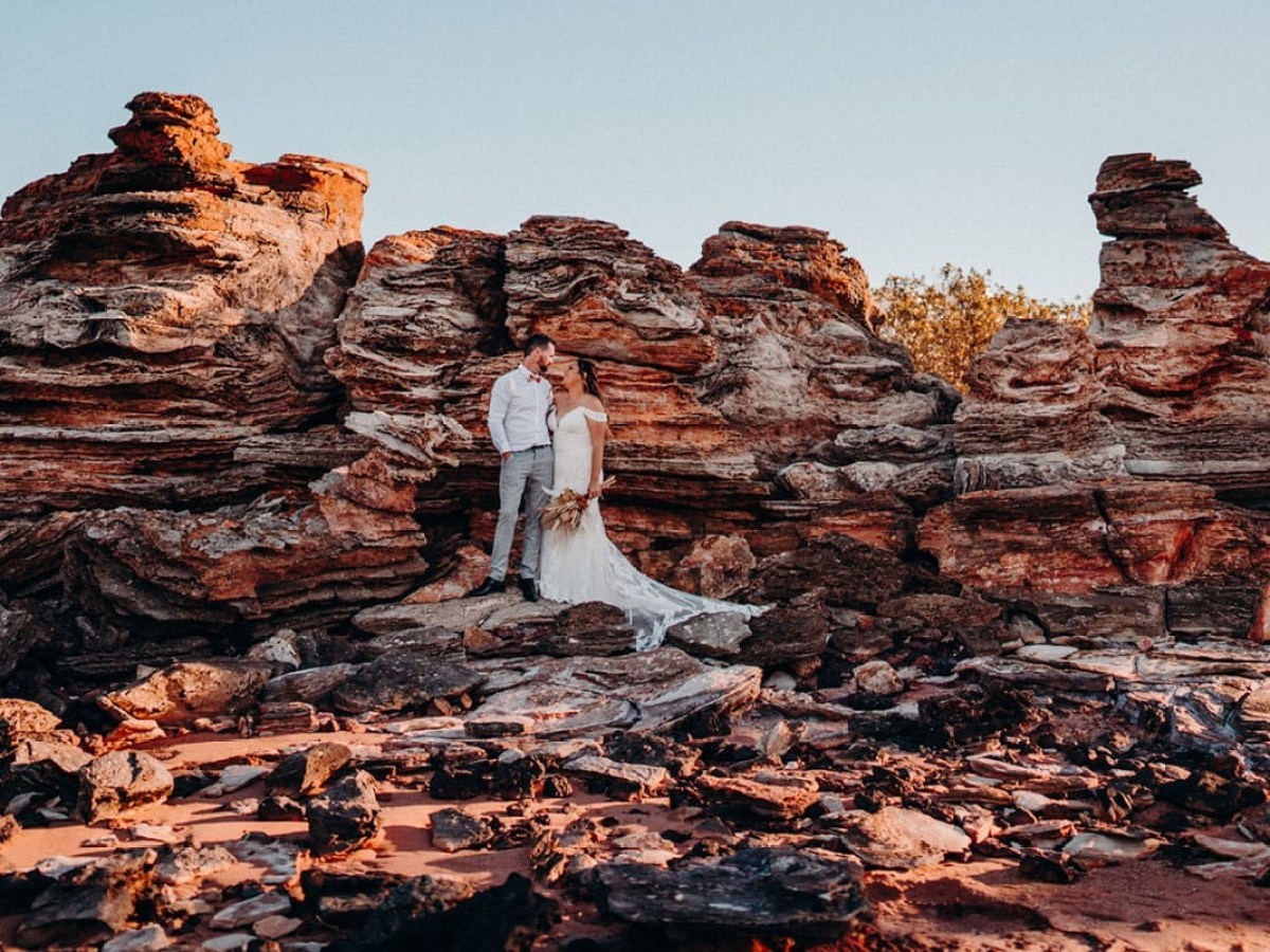 Couple in wedding attire stands by rugged, red rock formations under clear blue sky.