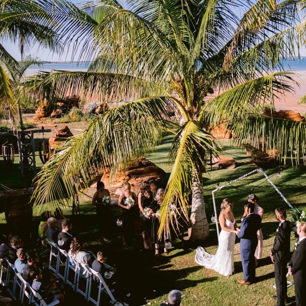 Outdoor wedding ceremony under palm trees by the beach with guests seated.