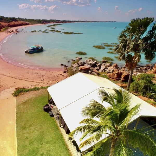 Aerial view of a coastal cabin with a palm tree, beach, and boat in turquoise waters.