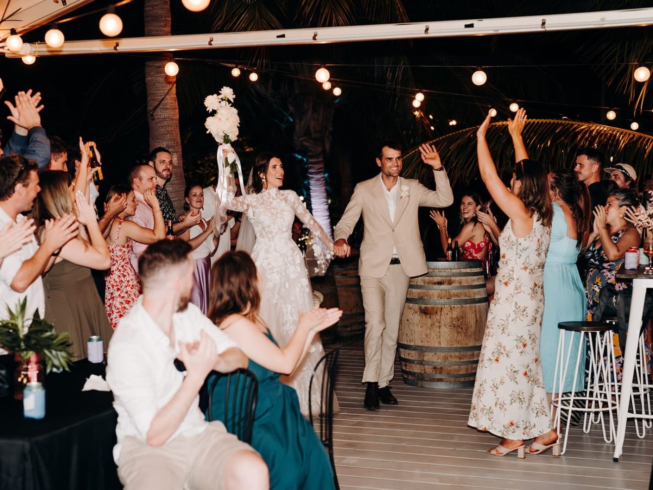 Couple in wedding attire entering reception area with guests cheering.