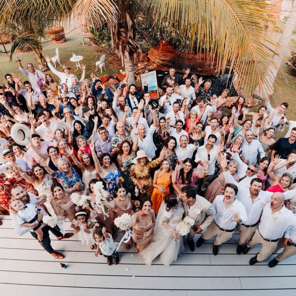 Aerial view of a large group of people at an outdoor event, smiling and waving under palm trees.