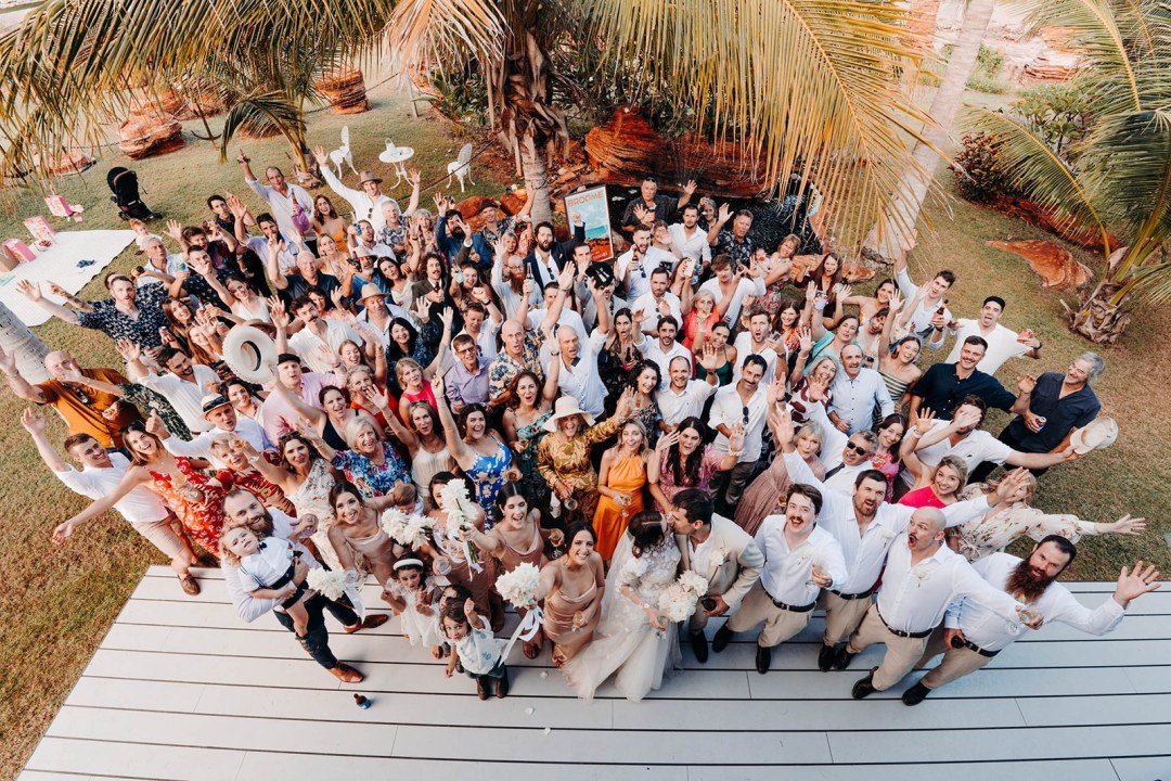 Aerial view of a large group of people at an outdoor event, smiling and waving under palm trees.