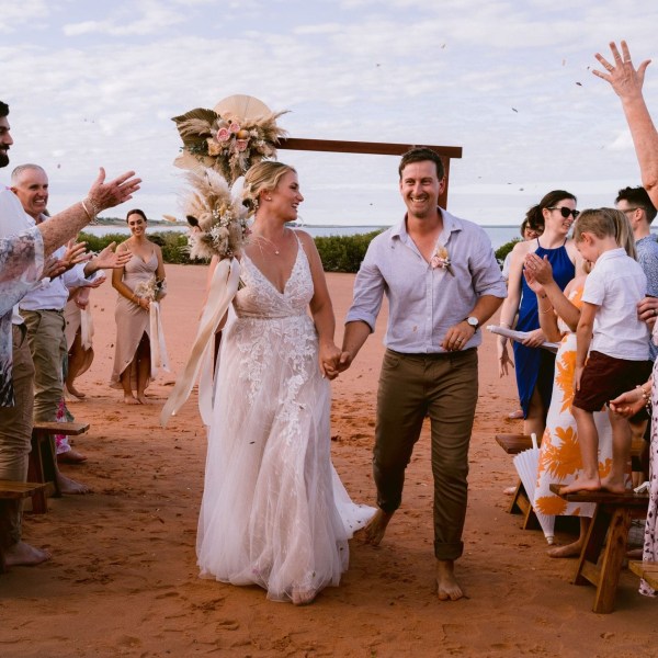 Bride and groom walking down sandy aisle, guests celebrating on beach.