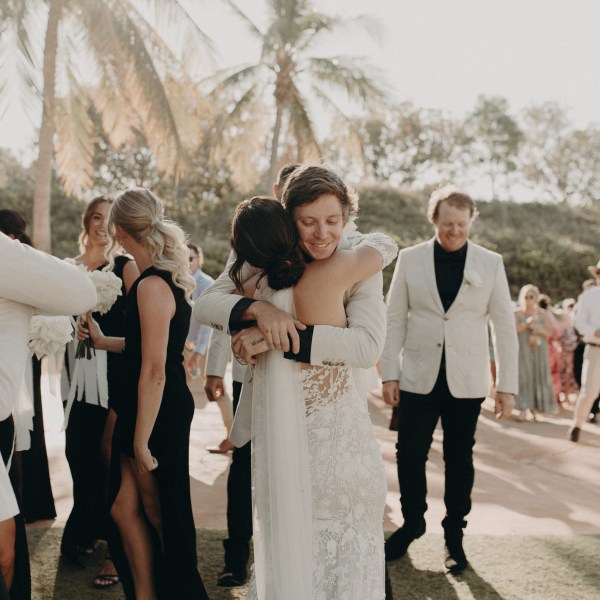 A couple hugs at an outdoor wedding with guests and palm trees in the background.