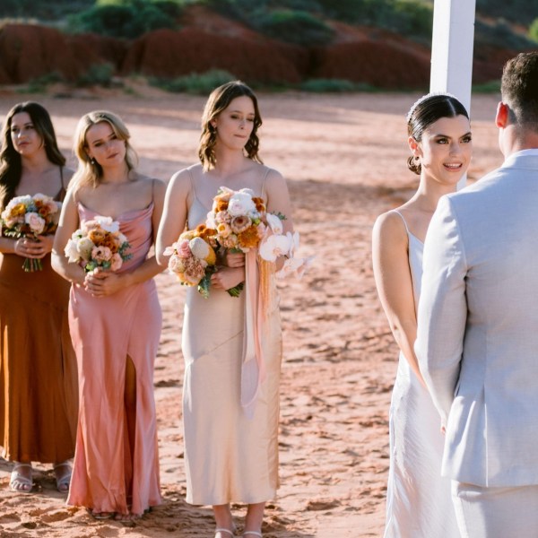 Bride and groom at outdoor ceremony, with bridesmaids holding flowers in the background.