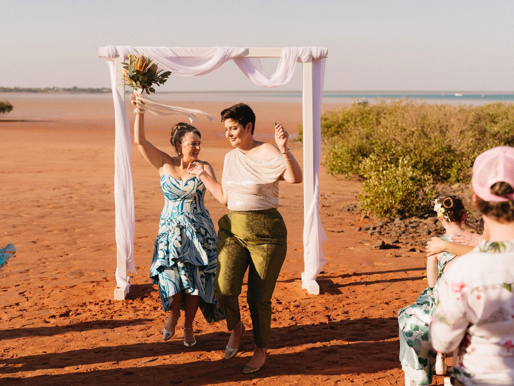 Two people joyfully exit a beach wedding arch, one holding a bouquet, with guests celebrating nearby.