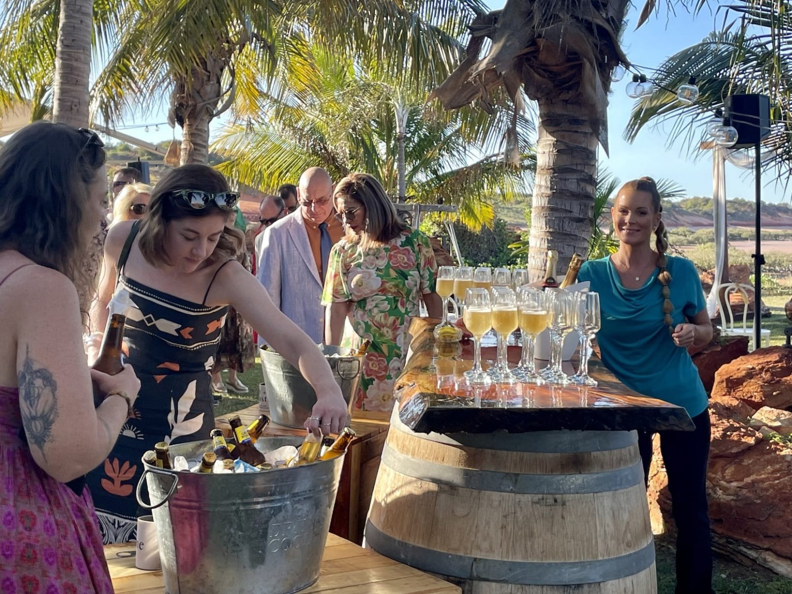People gathering around a champagne and beer station at an outdoor party with palm trees.