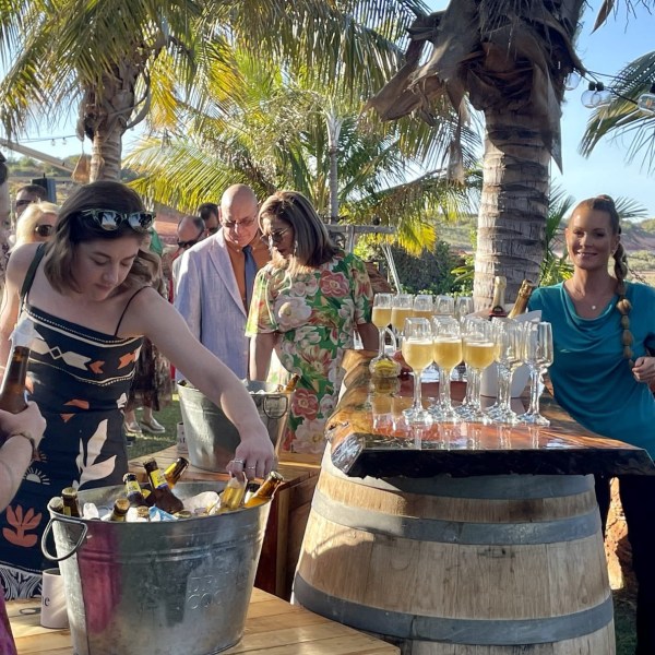 People gathering around a champagne and beer station at an outdoor party with palm trees.