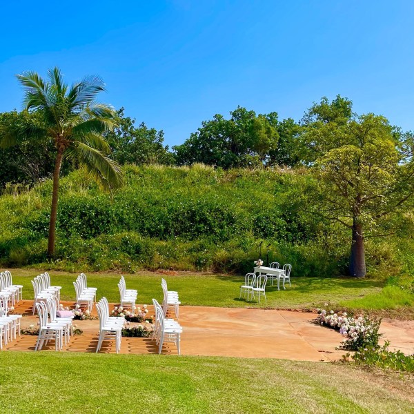 Outdoor wedding setup with white chairs, palm tree, and green hill background.