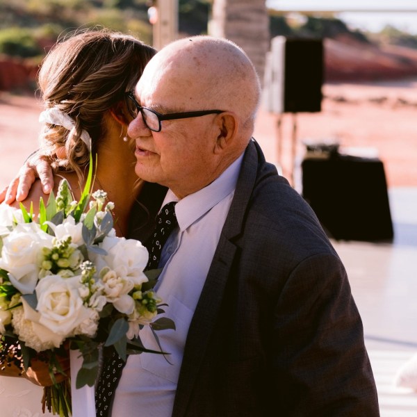 A bride holding flowers hugs an older man in glasses during an outdoor wedding.