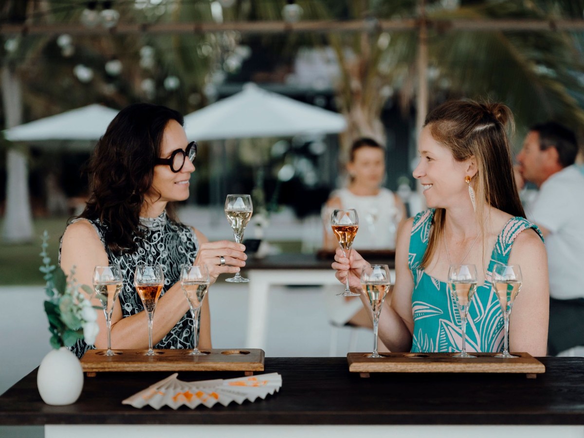 Two women sitting at a table outdoors toasting with rose wine glasses, with blurred people and umbrellas in the background.