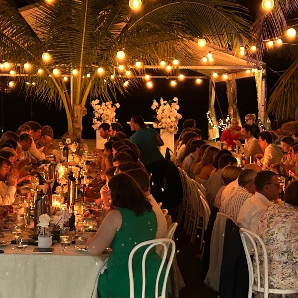 Outdoor dinner party with long tables, warm lighting, and guests under a tent and palm trees.