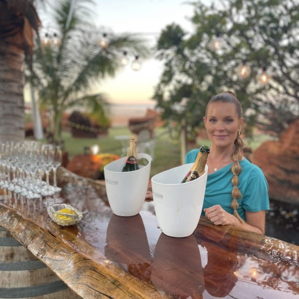 Woman behind outdoor bar with champagne in ice buckets and palm trees in background.