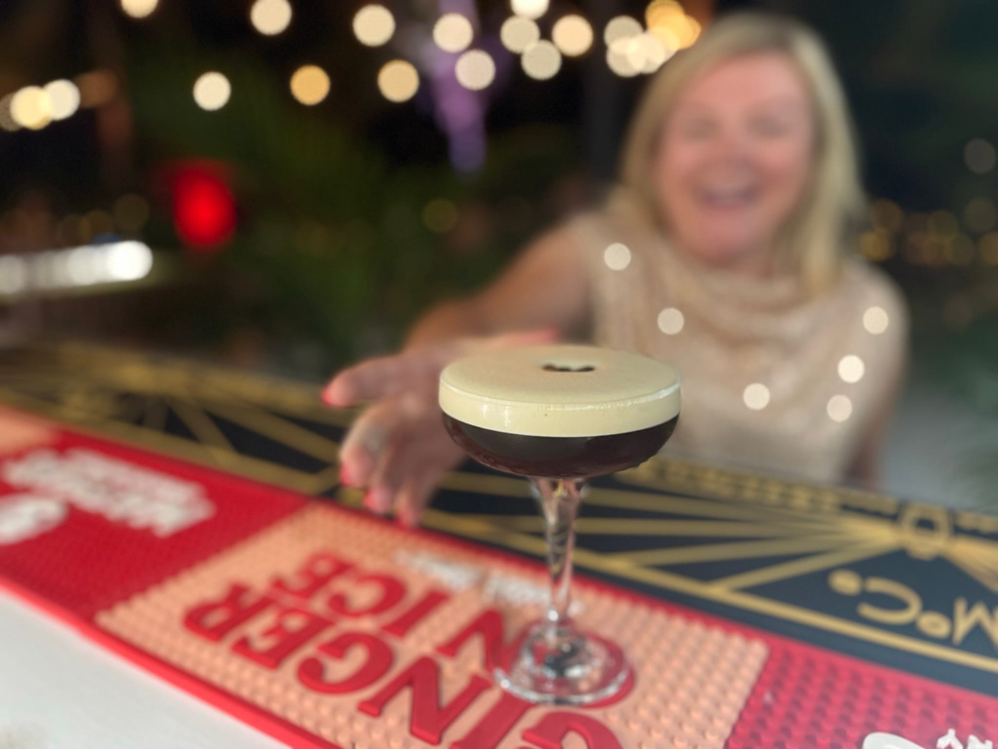 Woman reaching for a cocktail with a creamy top at a bar, blurred festive lights in background.